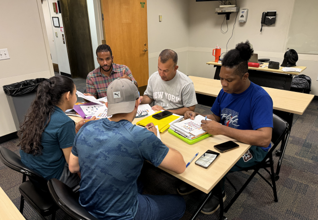 Adult students gather around a table with study materials, practicing their English language skills.